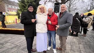 Vier Personen stehen in einem Park und lächeln für ein Foto. Eine Frau in einem weißen Kleid steht zwischen zwei Männern, einer hält ein Mikrofon. Hinter ihnen steht ein Lautsprecher und Bäume.