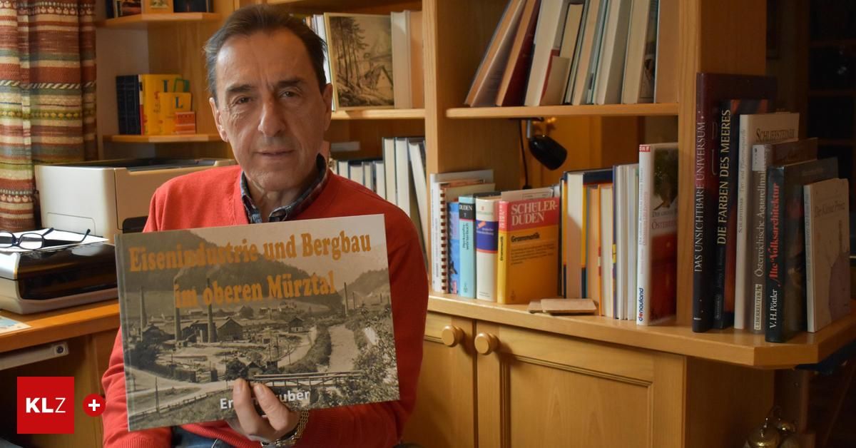 Ein älterer Mann in einem roten Pullover hält ein Buch mit dem Titel 'Bergbau und Bergbau im oberen Murztal'. Er steht vor einem Holzregal mit vielen Büchern.