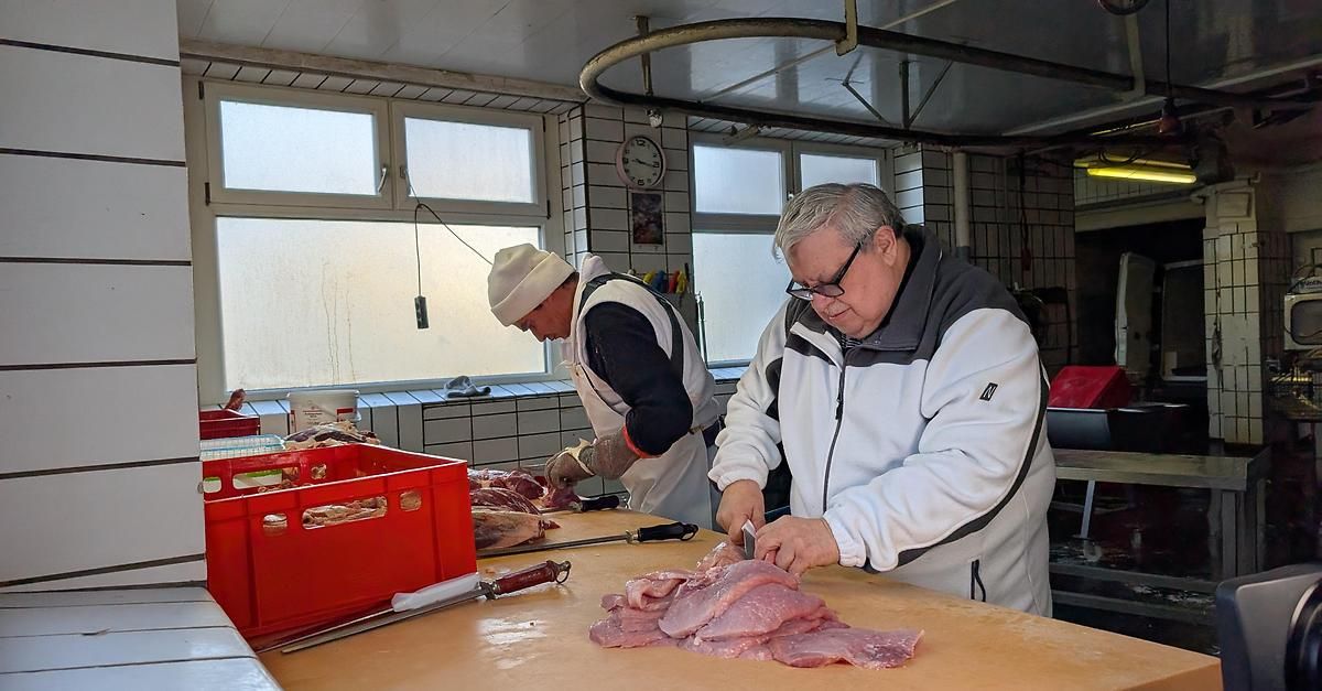 Zwei Männer in einem Metzgerladen schneiden Fleisch. Ein Mann trägt einen weißen Hut und eine weiße Schürze, während der andere eine Brille trägt. Ein roter Aufbewahrungskasten und ein Messer liegen auf dem Tisch. An der Wand hängt eine Uhr.