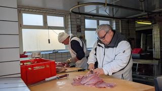 Zwei Männer in einem Metzgerladen schneiden Fleisch. Ein Mann trägt einen weißen Hut und eine weiße Schürze, während der andere eine Brille trägt. Ein roter Aufbewahrungskasten und ein Messer liegen auf dem Tisch. An der Wand hängt eine Uhr.