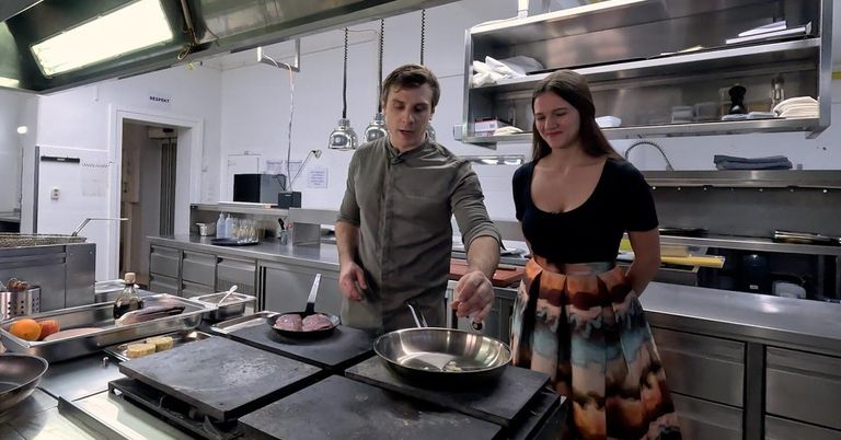 Ein Mann und eine Frau kochen in einer Küche. Der Mann benutzt einen Pfannenwender, um Essen in einer Bratpfanne zu wenden. Die Frau steht neben ihm und beobachtet.