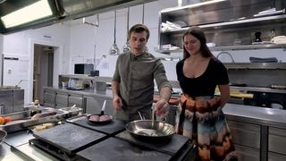 Ein Mann und eine Frau kochen in einer Küche. Der Mann benutzt einen Pfannenwender, um Essen in einer Bratpfanne zu wenden. Die Frau steht neben ihm und beobachtet.