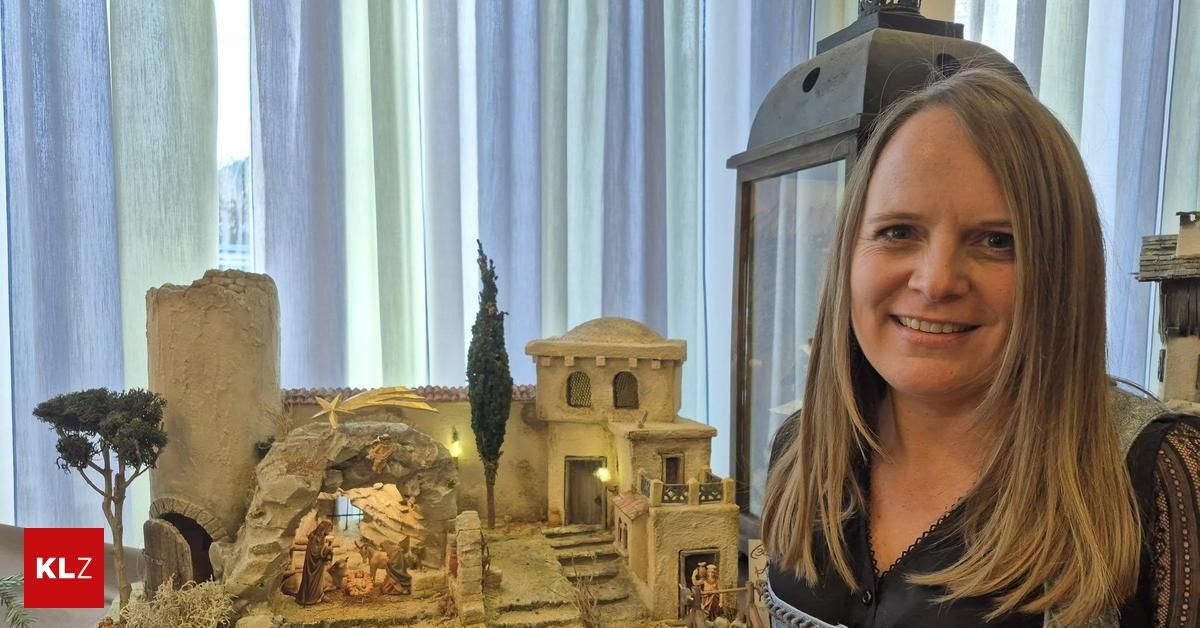 Eine Frau steht lächelnd vor einer Miniatur-Krippenszene mit einer Kirche, Stufen und einem Baum. Sie trägt ein schwarzes Oberteil und scheint sich in einem Raum mit einem weißen Vorhang zu befinden.