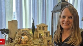 Eine Frau steht lächelnd vor einer Miniatur-Krippenszene mit einer Kirche, Stufen und einem Baum. Sie trägt ein schwarzes Oberteil und scheint sich in einem Raum mit einem weißen Vorhang zu befinden.