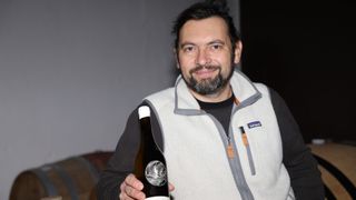 Ein Mann hält eine Flasche Wein in der Hand, trägt eine Jacke mit Logo und lächelt.