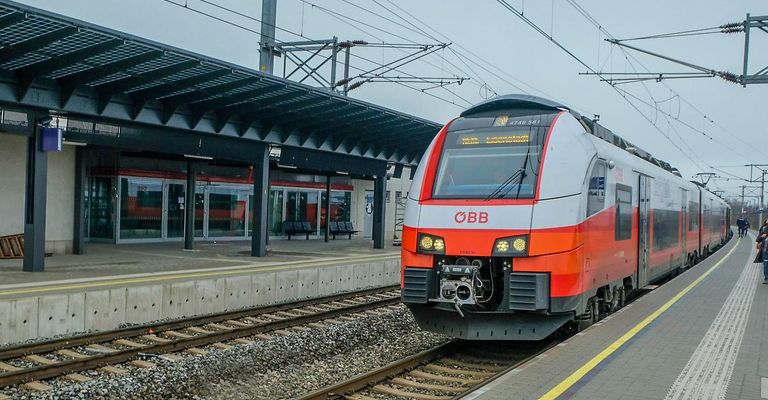 Ein orange-grauer Zug, gekennzeichnet durch OBB, steht auf einem Bahnsteig unter einem überhängenden Dach, mit Oberleitungen und Bänken im Sichtfeld.