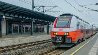 Ein orange-grauer Zug, gekennzeichnet durch OBB, steht auf einem Bahnsteig unter einem überhängenden Dach, mit Oberleitungen und Bänken im Sichtfeld.