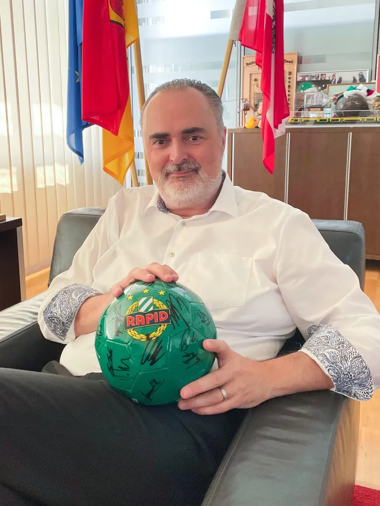 Ein Mann sitzt auf einem Stuhl und hält einen Fußball mit Unterschriften. Dahinter befinden sich Flaggen und ein hölzerner Schrank.