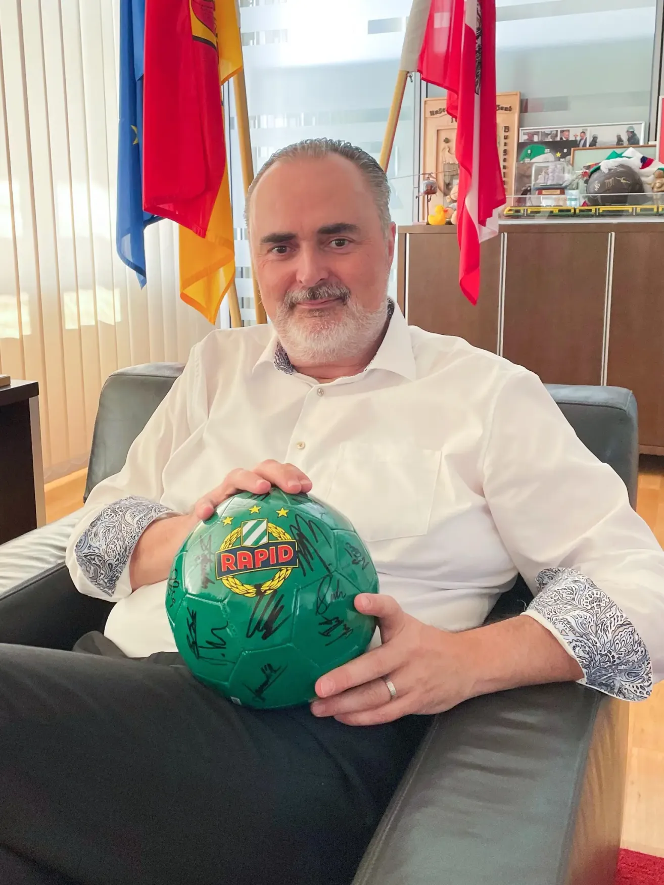 Ein Mann sitzt auf einem Stuhl und hält einen Fußball mit Unterschriften. Dahinter befinden sich Flaggen und ein hölzerner Schrank.