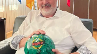 Ein Mann sitzt auf einem Stuhl und hält einen Fußball mit Unterschriften. Dahinter befinden sich Flaggen und ein hölzerner Schrank.