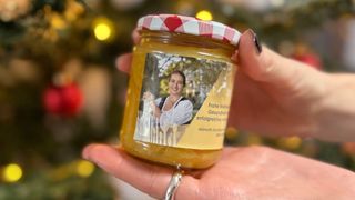 Eine Hand hält ein kleines Glas Honig mit einem festlichen Etikett, das eine Frau zeigt, vor dem Hintergrund eines Weihnachtsbaums.