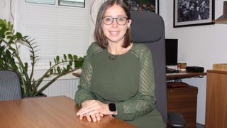 Eine Frau mit Brille und einem grünen Kleid sitzt lächelnd am Schreibtisch, mit einer Pflanze und einem Computer hinter ihr.