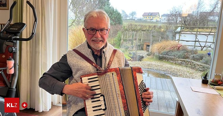 Ein älterer Mann spielt Akkordeon in einem Zimmer mit einem Glasfenster, das eine malerische Aussicht auf ein Haus und Bäume zeigt.