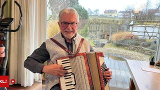 Ein älterer Mann spielt Akkordeon in einem Zimmer mit einem Glasfenster, das eine malerische Aussicht auf ein Haus und Bäume zeigt.