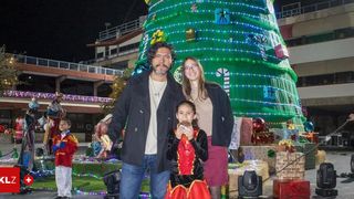 Ein Mann, eine Frau und ein junges Mädchen stehen nachts vor einem großen Weihnachtsbaum. Der Mann trägt eine Halskette und die Frau eine Brille. Das Mädchen ist in einem rot-schwarzen Kostüm gekleidet. Sie alle lächeln und scheinen für ein Foto zu posieren. Hinter ihnen befinden sich weitere Weihnachtsdekorationen und ein kleines Kind in einem roten Kostüm.