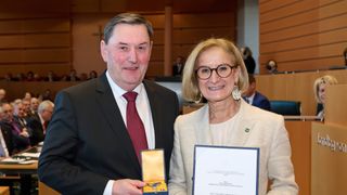 Ein Mann und eine Frau in formeller Kleidung lächeln und halten Auszeichnungen, stehen in einem Konferenzsaal.