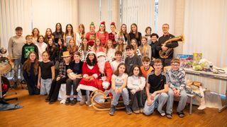 Eine Gruppe von Menschen posiert für ein Weihnachtsfoto mit dem Weihnachtsmann und einem Baby. Sie tragen Winterkleidung und einige haben Mützen. Ein Mann spielt ein Blasinstrument.