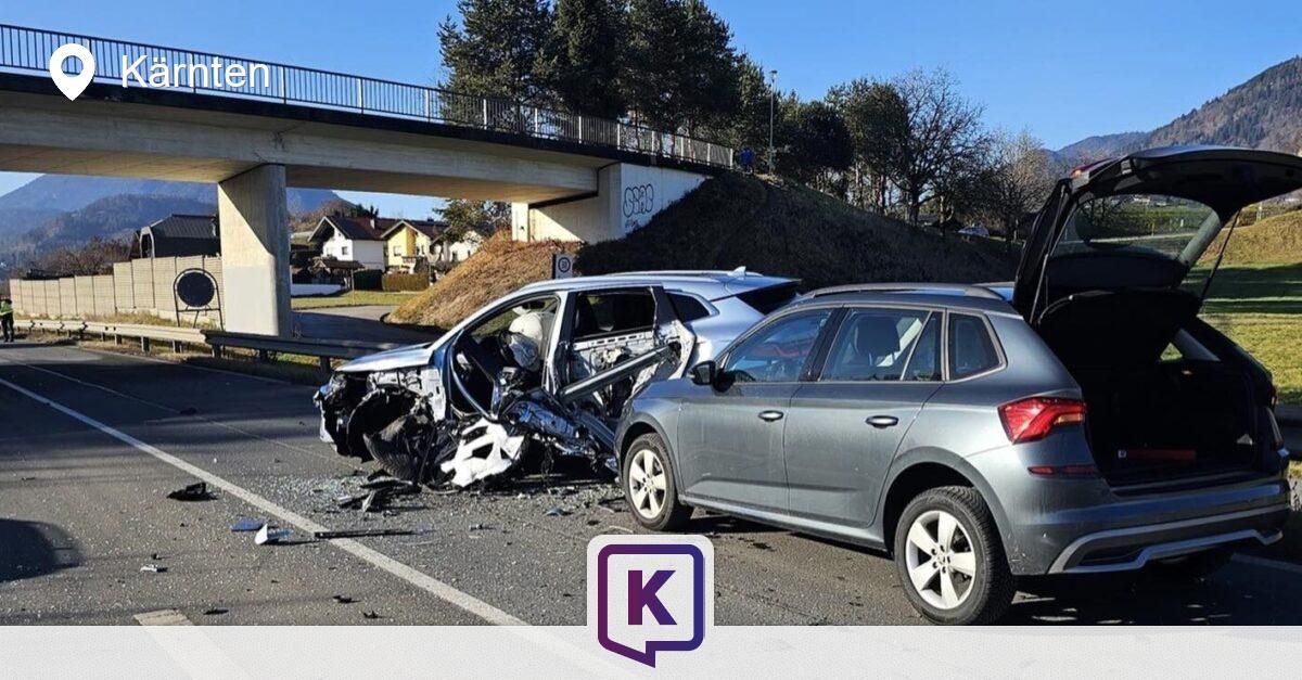 Zwei Autos sind auf einer Straße zerstört, wobei ein Auto die vordere Seite zerschmettert hat und das andere Auto die hintere Tür offen hat. Eine Brücke und ein Hang mit Bäumen sind im Hintergrund.