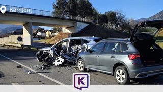 Zwei Autos sind auf einer Straße zerstört, wobei ein Auto die vordere Seite zerschmettert hat und das andere Auto die hintere Tür offen hat. Eine Brücke und ein Hang mit Bäumen sind im Hintergrund.