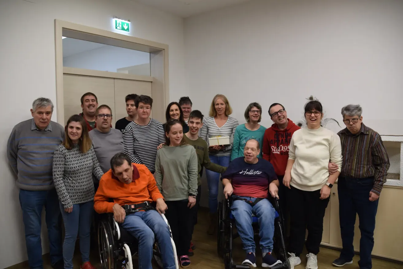 Eine Gruppe von Menschen steht in einem Raum zusammen. Einige sitzen in Rollstühlen, andere stehen. Sie lächeln und posieren für ein Foto. Der Raum hat Holzböden und weiße Wände.