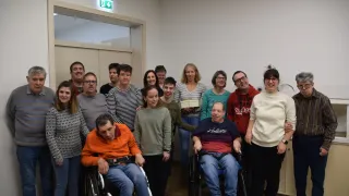 Eine Gruppe von Menschen steht in einem Raum zusammen. Einige sitzen in Rollstühlen, andere stehen. Sie lächeln und posieren für ein Foto. Der Raum hat Holzböden und weiße Wände.