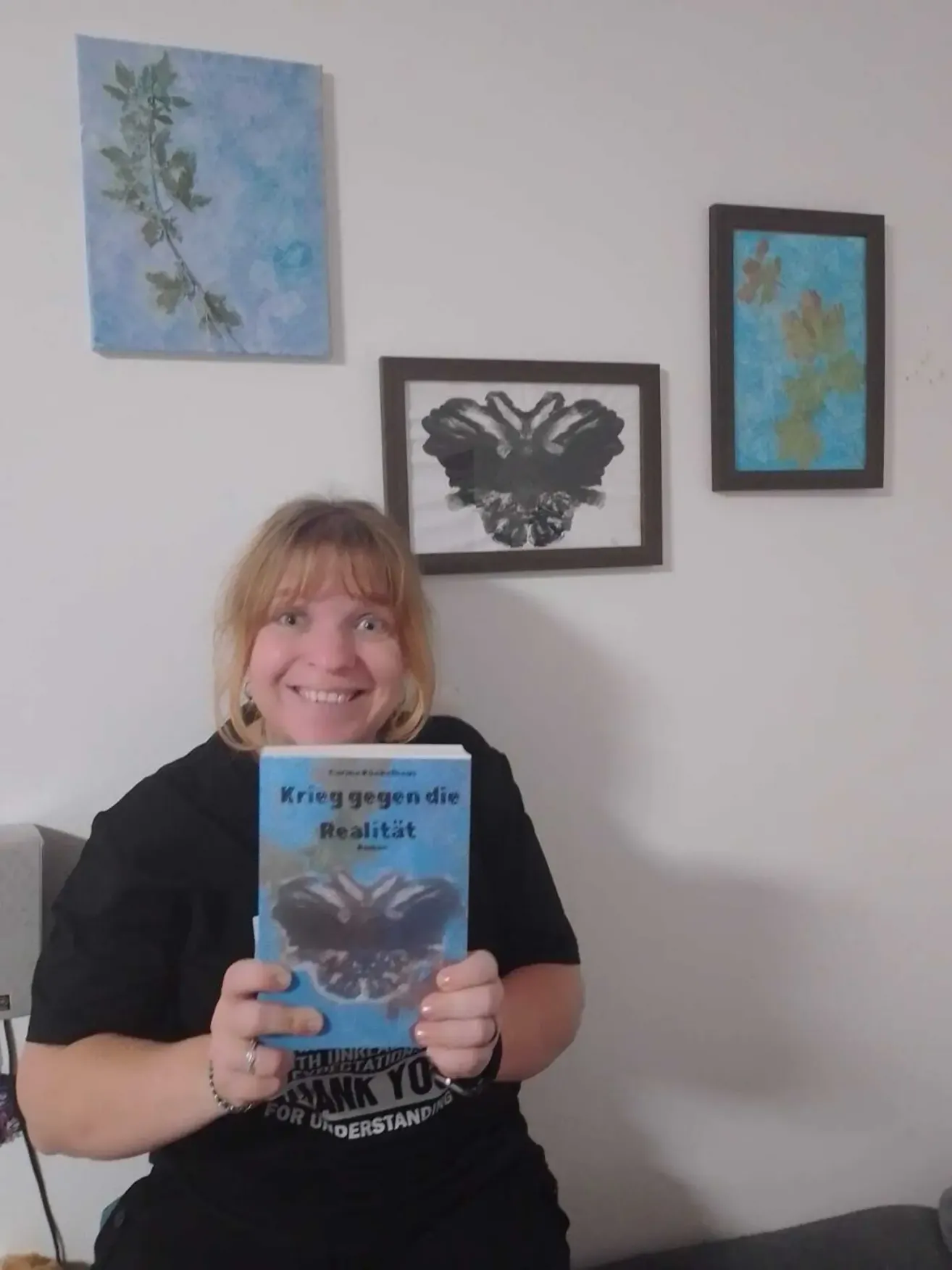 Eine Frau hält ein Buch mit dem Titel 'Krieg gegen die Realität' und lächelt. Sie steht vor einer weißen Wand mit drei gerahmten Bildern.