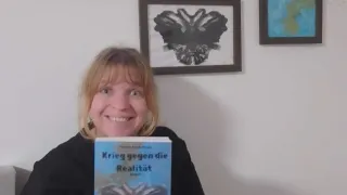 Eine Frau hält ein Buch mit dem Titel 'Krieg gegen die Realität' und lächelt. Sie steht vor einer weißen Wand mit drei gerahmten Bildern.