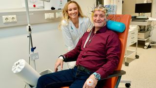 Ein Mann im Krankenhausstuhl mit EEG-Kappe lächelt, während eine Frau in einem Laborkittel hinter ihm steht, beide schauen in die Kamera.