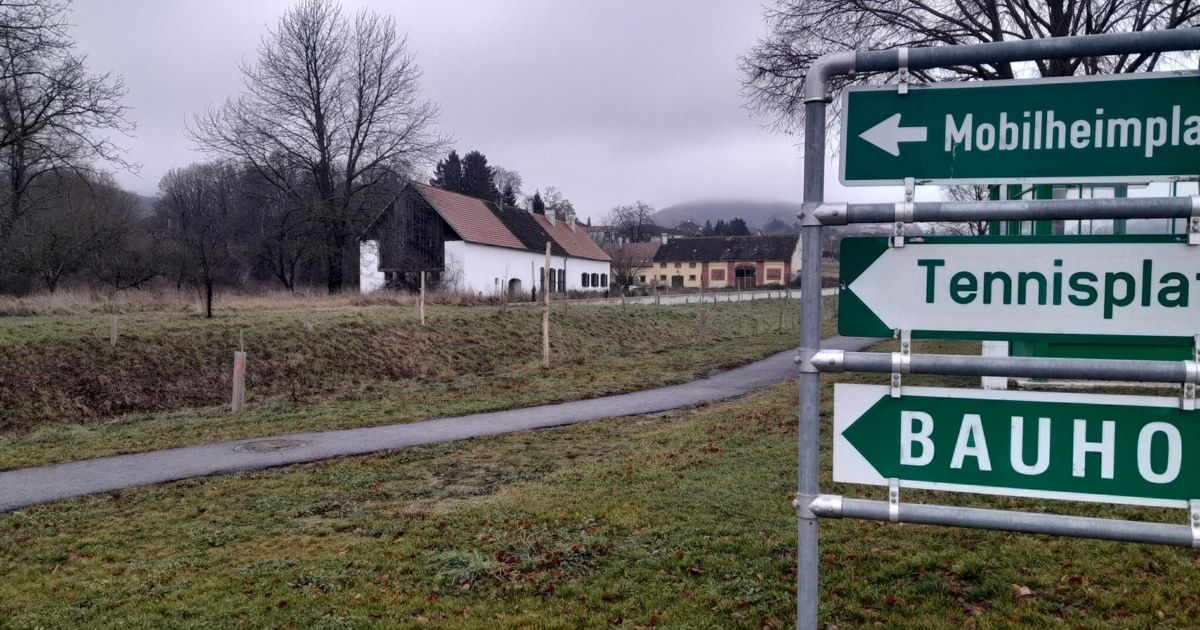 Ein Wegweiser mit grünen Pfeilen zeigt Richtungen zu M, Te und B an. Im Hintergrund befindet sich ein Haus mit rotem Dach und einem Weg, der dorthin führt.