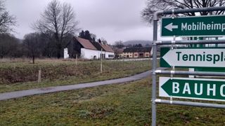 Ein Wegweiser mit grünen Pfeilen zeigt Richtungen zu M, Te und B an. Im Hintergrund befindet sich ein Haus mit rotem Dach und einem Weg, der dorthin führt.