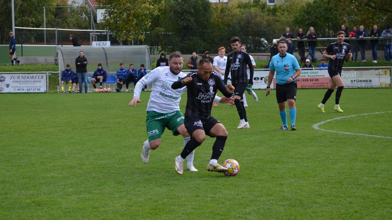 Fußballspieler konkurrieren auf einem Feld, wobei zwei Spieler im Fokus stehen, einer in Weiß und einer in Schwarz, die um den Ball kämpfen. Zuschauer sitzen auf blauen Stühlen hinter einem Zaun.