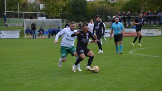 Fußballspieler konkurrieren auf einem Feld, wobei zwei Spieler im Fokus stehen, einer in Weiß und einer in Schwarz, die um den Ball kämpfen. Zuschauer sitzen auf blauen Stühlen hinter einem Zaun.