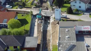 Ein Luftbild einer Nachbarschaft mit mehreren Häusern und Gebäuden im Bau. Ein gelber Bagger arbeitet auf einer Baustelle. Das Gebiet hat grünen Rasen, Bäume und eine unbefestigte Straße.