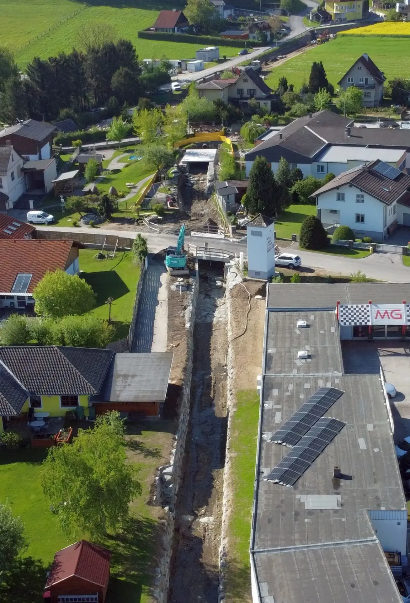 Ein Luftbild einer Nachbarschaft mit mehreren Häusern und Gebäuden im Bau. Ein gelber Bagger arbeitet auf einer Baustelle. Das Gebiet hat grünen Rasen, Bäume und eine unbefestigte Straße.