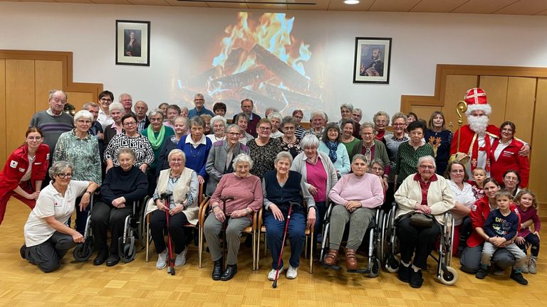 Eine Gruppe älterer Menschen in Rollstühlen und Stühlen, die für ein Foto vor einer Feuerprojektion an der Wand hinter ihnen posieren.