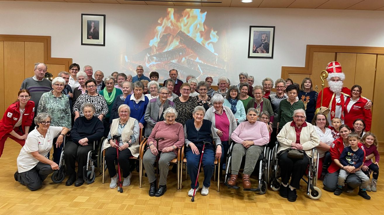 Eine Gruppe älterer Menschen in Rollstühlen und Stühlen, die für ein Foto vor einer Feuerprojektion an der Wand hinter ihnen posieren.