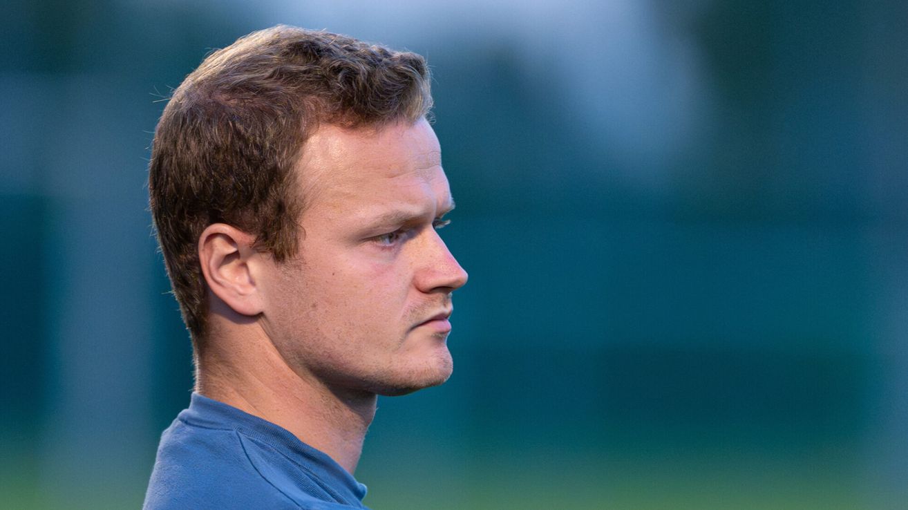 Ein Mann mit kurzem, braunem Haar und einem blauen T-Shirt schaut mit ernster Miene nach rechts. Der Hintergrund ist unscharf, was auf ein Feld hindeutet.