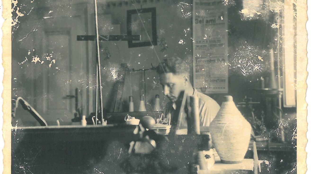 Ein Schwarz-Weiß-Foto einer Frau in einer Werkstatt, konzentriert auf ein Töpferprojekt. Sie sitzt mit Werkzeugen und Töpferstücken um sie herum.