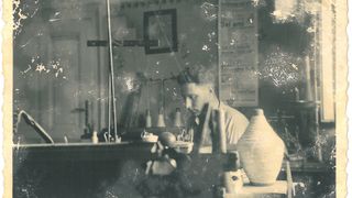 Ein Schwarz-Weiß-Foto einer Frau in einer Werkstatt, konzentriert auf ein Töpferprojekt. Sie sitzt mit Werkzeugen und Töpferstücken um sie herum.