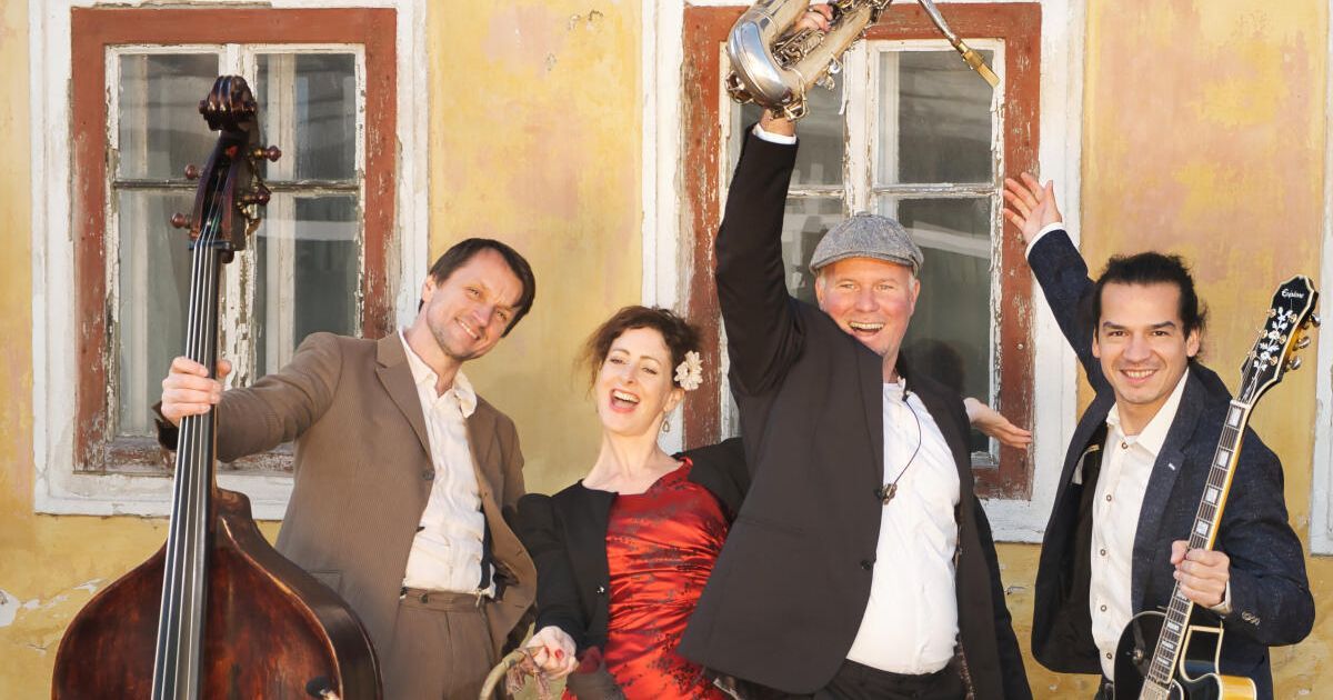 Eine Gruppe von Musikern posiert für ein Foto, wobei einer einen Saxophon und der andere einen Cello hält, beide lächeln fröhlich vor einem gelben Gebäude.