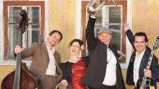 Eine Gruppe von Musikern posiert für ein Foto, wobei einer einen Saxophon und der andere einen Cello hält, beide lächeln fröhlich vor einem gelben Gebäude.