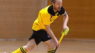 Ein Mann spielt Feldhockey in einer Indoor-Arena und trägt ein gelbes und schwarzes Trikot mit einem Logo auf der Brust.