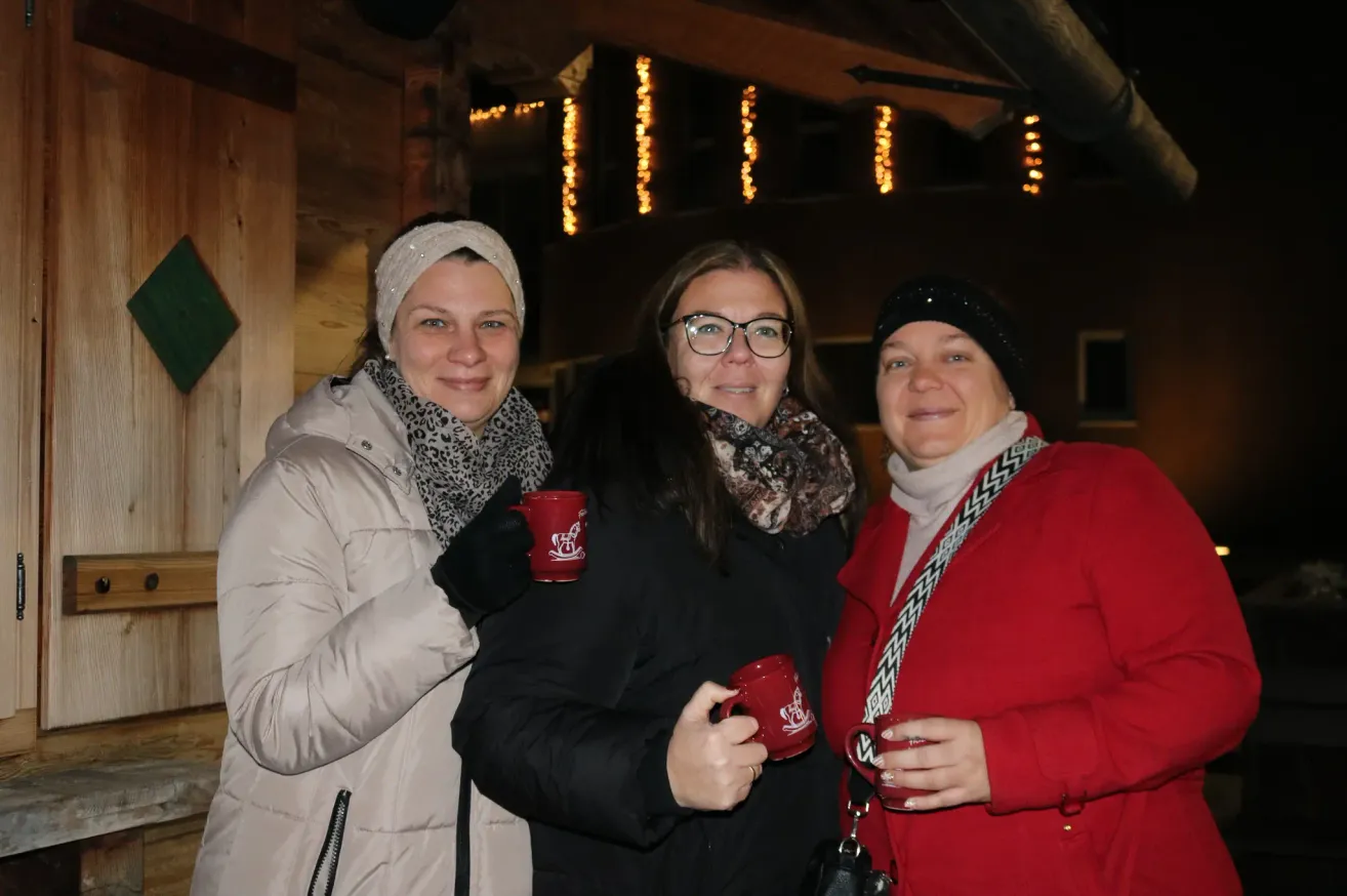 Drei Frauen in Wintermänteln und Mützen lächeln für die Kamera. Jede hält einen roten Becher mit Logo. Dahinter ist ein Gebäude mit Lichterketten geschmückt.