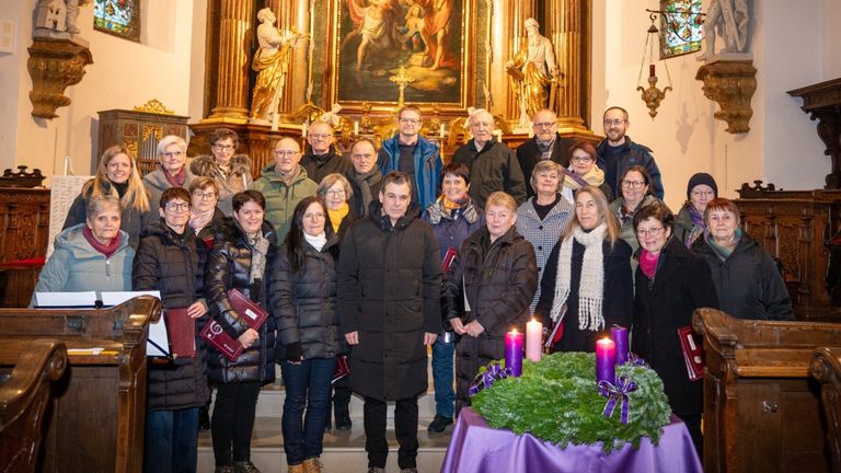 Eine Gruppe von Menschen steht in einer Kirche, warm gekleidet für den Winter. Sie sind um einen Tisch mit einem Kranz und Kerzen versammelt. Hinter ihnen schmücken Statuen und ein großes Gemälde den Altar.