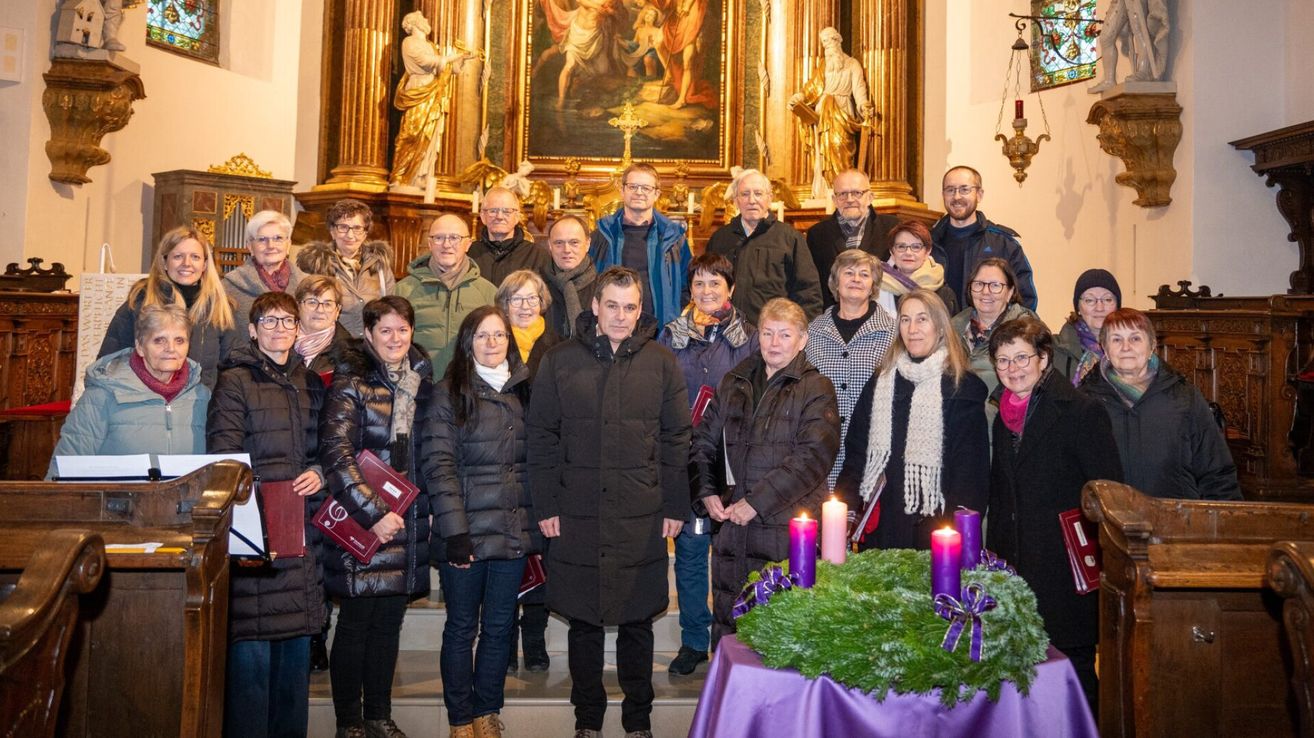 Eine Gruppe von Menschen steht in einer Kirche, warm gekleidet für den Winter. Sie sind um einen Tisch mit einem Kranz und Kerzen versammelt. Hinter ihnen schmücken Statuen und ein großes Gemälde den Altar.