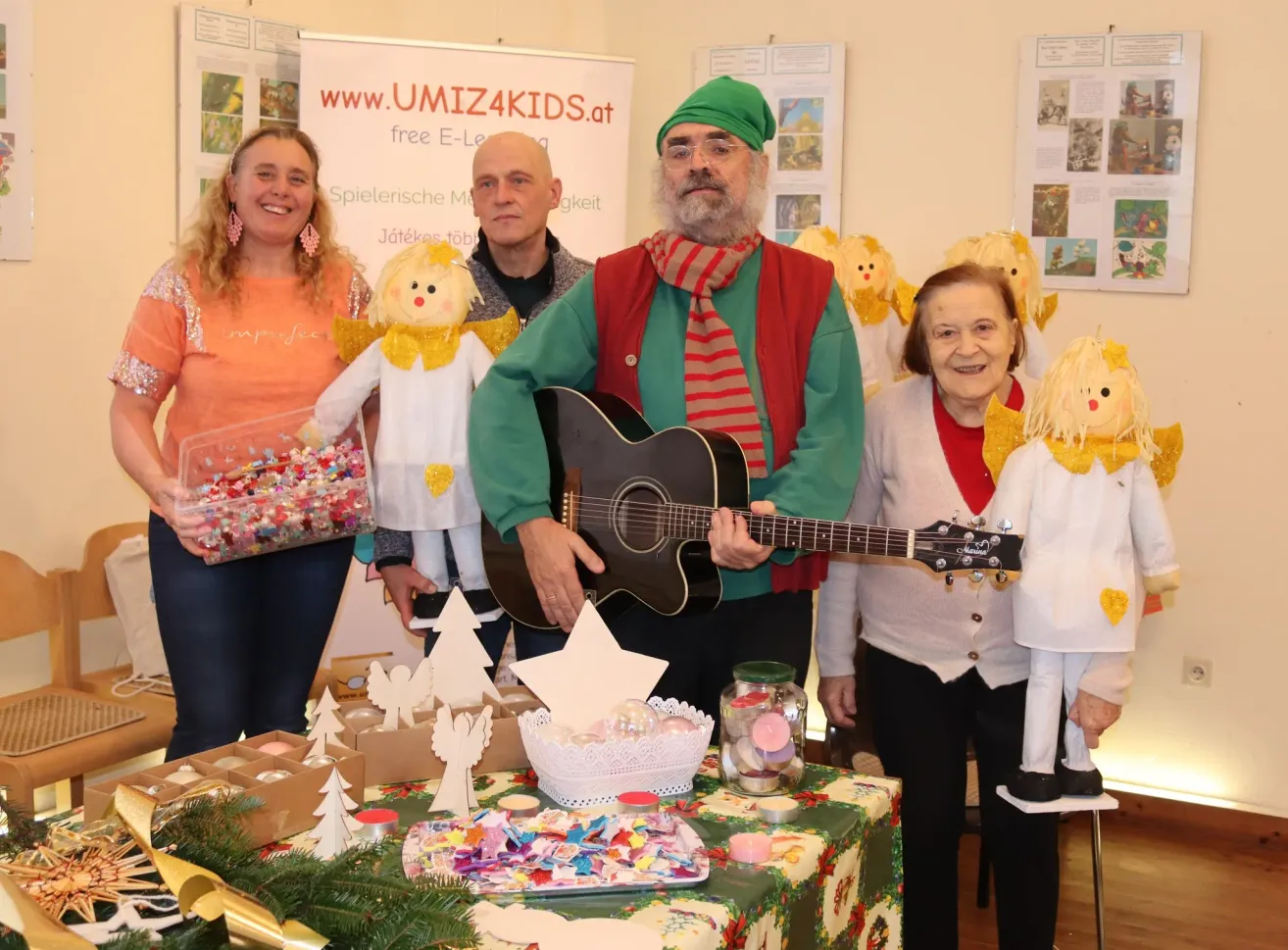 Fünf Menschen posieren für ein Foto, darunter eine Frau mit grünem Hut und Gitarre, eine Frau in Orange, die eine Schachtel mit Süßigkeiten hält, und ein Mann mit rotem Schal. Es gibt Weihnachtsdekorationen auf dem Tisch.