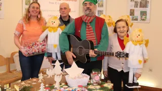 Fünf Menschen posieren für ein Foto, darunter eine Frau mit grünem Hut und Gitarre, eine Frau in Orange, die eine Schachtel mit Süßigkeiten hält, und ein Mann mit rotem Schal. Es gibt Weihnachtsdekorationen auf dem Tisch.