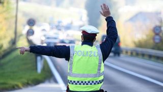 Ein Verkehrspolizist leitet den Verkehr auf einer Autobahn. Er trägt eine reflektierende Weste und einen Hut, und sein rechter Arm ist erhoben.
