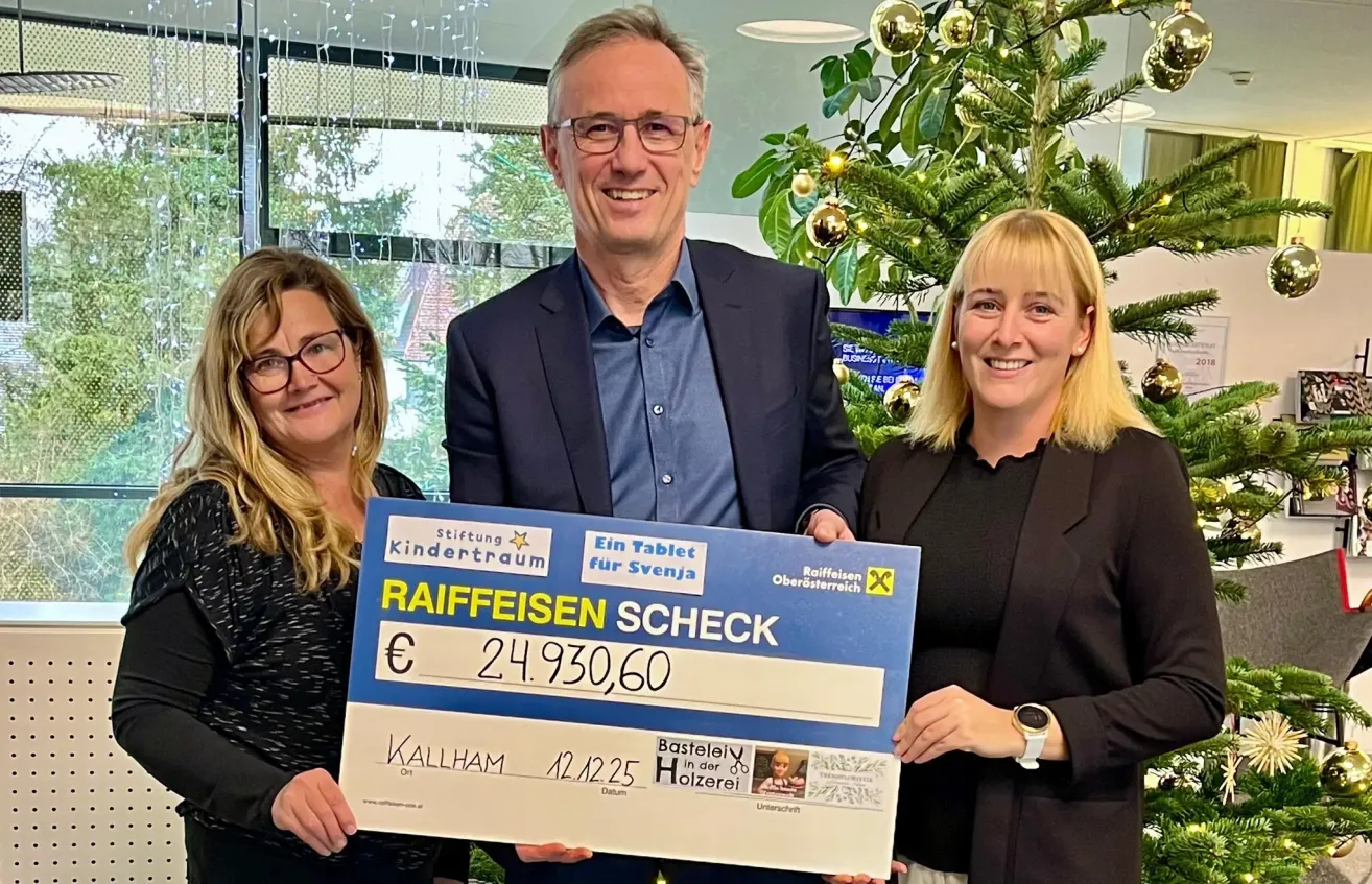 Drei Personen stehen nebeneinander und halten einen großen Scheck mit den Worten Raiffeisen-Scheck, einer Bank in Österreich. Der Scheck ist für 24.930,60 Euro. Der Mann in der Mitte trägt eine Brille und einen Anzug, während die Frauen auf beiden Seiten von ihm schwarz gekleidet sind. Hinter ihnen steht ein Weihnachtsbaum und ein Fenster mit Blick auf Bäume.