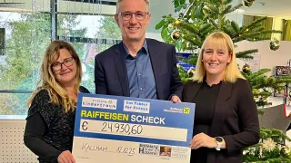 Drei Personen stehen nebeneinander und halten einen großen Scheck mit den Worten Raiffeisen-Scheck, einer Bank in Österreich. Der Scheck ist für 24.930,60 Euro. Der Mann in der Mitte trägt eine Brille und einen Anzug, während die Frauen auf beiden Seiten von ihm schwarz gekleidet sind. Hinter ihnen steht ein Weihnachtsbaum und ein Fenster mit Blick auf Bäume.